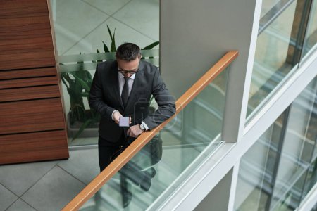 Foto de Joven abogado o corredor en ropa formal usando teléfono inteligente mientras está de pie junto a la barandilla en uno de los grandes múltiples pisos moderno centro de oficinas - Imagen libre de derechos