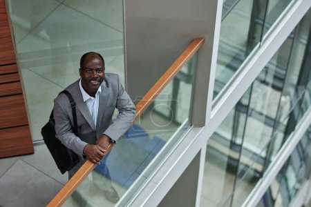 Téléchargez les photos : Homme d'affaires afro-américain mature en costume gris regardant la caméra avec le sourire tout en étant debout par une rampe transparente au centre du bureau - en image libre de droit