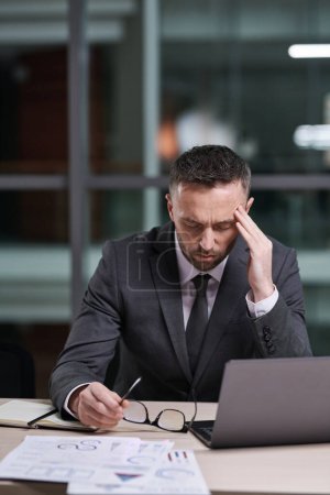 Photo for Overworked and tired businessman in formalwear sitting by desk, bending over laptop and touching temple while trying to concentrate - Royalty Free Image