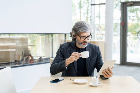 Téléchargez les photos : Barbu économiste mature ou banquier en costume et lunettes d'analyse des données financières en ligne dans la tablette par tasse de café dans le restaurant moderne - en image libre de droit