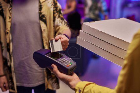 Téléchargez les photos : Jeune homme en tenue décontractée payant par carte de crédit pour la pizza commandée emballée dans des boîtes carrées en carton tenues par le livreur en uniforme - en image libre de droit