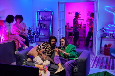 Foto de Joven pareja intercultural mirando a través de nuevas fotos en el teléfono móvil mientras se sienta en el sofá en la fiesta en casa contra sus amigos - Imagen libre de derechos