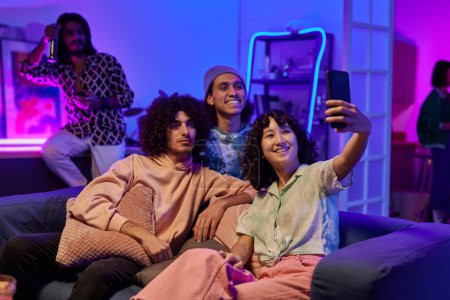 Foto de Felices amigos interculturales jóvenes en ropa de casualwear tomar selfie en la fiesta en casa mientras se relaja en el cómodo sofá en la sala de estar - Imagen libre de derechos