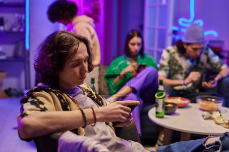 Foto de Joven hombre descansado en ropa de casualwear sentado en sillón contra sus amigos en la sala de estar iluminado por la luz de neón en la fiesta en casa y el uso de teléfono móvil - Imagen libre de derechos