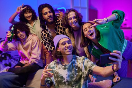 Group of young cheerful intercultural friends in casualwear taking selfie at home party while standing in living room lit by neon light