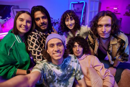 Foto de SIx jóvenes interculturales hombres y mujeres posando para selfie delante de la cámara mientras disfruta de la fiesta en casa en la sala de estar iluminada por la luz de neón - Imagen libre de derechos