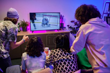 Téléchargez les photos : Vue arrière d'amis assis devant l'écran de télévision et regardant la diffusion du match de hockey dans le salon éclairé par la lumière du néon à loisir - en image libre de droit