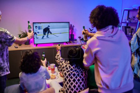 Foto de Respaldos de amigos jóvenes con botellas de cerveza reunidos frente al televisor celebrando la victoria de su equipo de hockey favorito en la fiesta en casa - Imagen libre de derechos