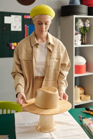 Photo for Young creative female designer of hats preparing wooden workpiece or mannequin on workplace while working over new models - Royalty Free Image