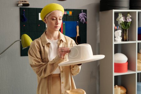 Téléchargez les photos : Jeune femme propriétaire sérieuse de la boutique artisanale et designer de la marque de mode créant un nouveau chapeau en feutre tout en se tenant près du lieu de travail devant la caméra - en image libre de droit