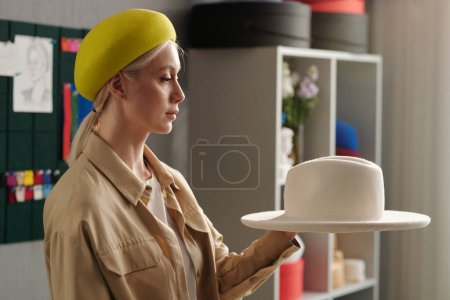 Photo for Young female stylist or owner of new fashion brand holding white hat and looking at trendy item from seasonal collection of accessories - Royalty Free Image