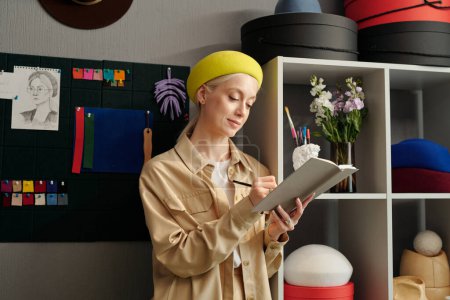 Photo for Young blond woman with notebook and pencil making notes or drawing sketches of new fashion items against wall with textile samples - Royalty Free Image