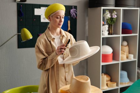 Téléchargez les photos : Jeune femme créative en béret jaune et casual chic debout en studio de mode et de nettoyage nouveau chapeau en feutre blanc avec brosse - en image libre de droit