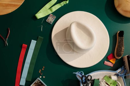 Téléchargez les photos : Vue du dessus du chapeau en feutre blanc parmi une variété d'échantillons textiles, broches dans une petite boîte, brosse pour nettoyer les nouveaux articles et autres fournitures sur la table - en image libre de droit