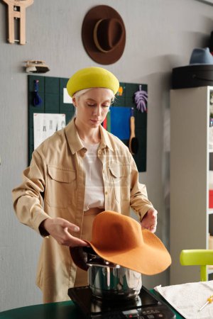 Foto de Fabricante de sombreros creativo joven en ropa casual inteligente de pie por el lugar de trabajo en el estudio de diseño y humeante nuevo artículo sobre la cacerola con agua hirviendo - Imagen libre de derechos