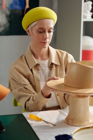 Téléchargez les photos : Jeune femme blonde mettant pièce en bois sur tige épaisse avant de le fixer tout en étant assis par le lieu de travail et la création de nouveaux chapeaux - en image libre de droit