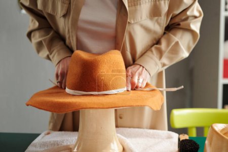 Photo for Close-up of young creative female artisan fixing white decorative shoelace around new brown handmade felt hat while decorating it before selling - Royalty Free Image