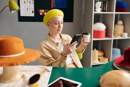 Foto de Joven artesana descansada usando teléfono inteligente y tomando café en el descanso en medio del día de trabajo mientras está sentada en un estudio o tienda de artesanía - Imagen libre de derechos