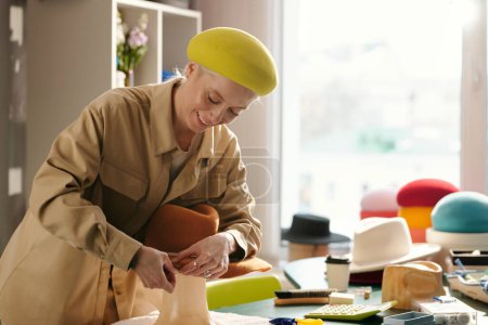 Téléchargez les photos : Sourire artisan féminin plier bords du bord du chapeau tout en le fixant avec des broches sur la pièce à usiner pendant le travail sur le nouvel article pour la vente dans l'atelier d'artisanat - en image libre de droit