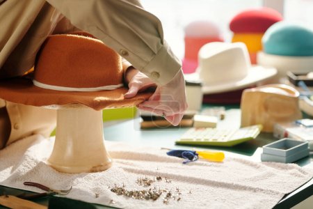 Téléchargez les photos : Gros plan du jeune artisan épinglant les bords du nouveau chapeau panama en feutre brun sur la pièce tout en restant près de la table avec des fournitures en studio - en image libre de droit