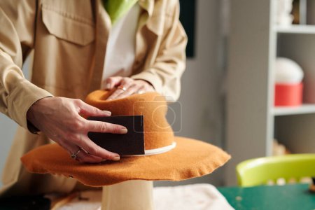 Photo for Hand of young craftswoman with polishing tool taking care of felt panama hat of brown color while creating new item for sale in craft shop - Royalty Free Image