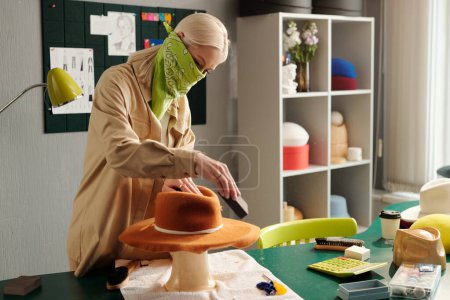 Photo for Young blond woman with protective bandana on face polishing brown felt hat with special tool while bending over workplace with supplies - Royalty Free Image