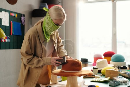Foto de Mujer creativa joven con bandana en la cara pulido nuevo sombrero de fieltro hecho a mano en el equipo especial mientras trabaja en la tienda de artesanía o estudio - Imagen libre de derechos