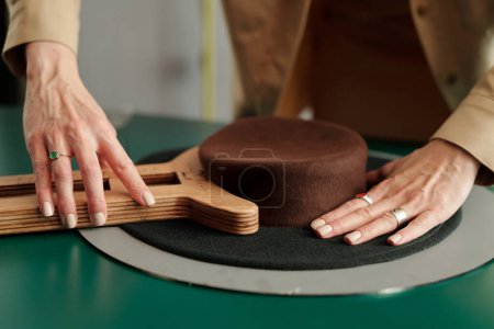 Foto de Manos de joven diseñadora formando forma de sombrero de cerdo de fieltro marrón con herramienta de madera por lugar de trabajo en estudio o pequeña tienda de artesanía - Imagen libre de derechos