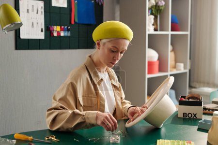Foto de Joven mujer creativa seria fijando cinta textil dentro de fieltro blanco sombrero panama mientras está sentado por el lugar de trabajo en la tienda de artesanía - Imagen libre de derechos
