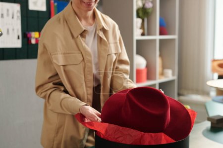 Téléchargez les photos : Jeune propriétaire de magasin de coiffure artisanale emballant un nouveau chapeau panama en feutre créé individuellement pour le client dans une boîte ronde noire avant l'emballage - en image libre de droit