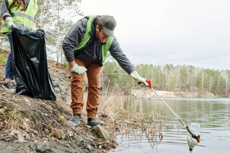 Foto de Hombre con palillo de recogida alcanzando para botella de plástico sucio en el lago - Imagen libre de derechos