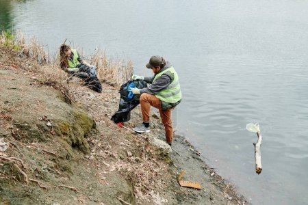 Téléchargez les photos : Couple nettoyage rive du lac et de mettre les ordures dans des sacs en plastique - en image libre de droit