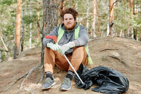 Téléchargez les photos : Volontaire barbu aux cheveux roux se reposant après avoir travaillé dans la forêt et ramassé des ordures - en image libre de droit