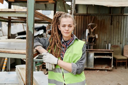 Photo for Portrait of positve female volunteer in neon vest and gloves standing at scrapyard and smiling at camera - Royalty Free Image