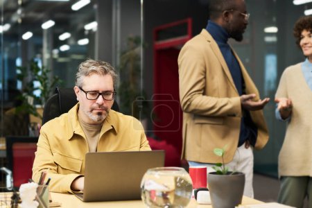 Foto de Serios hombres de negocios maduros en red delante de la computadora portátil mientras se sienta junto al escritorio en contra de dos jóvenes colegas interculturales que tienen discusión - Imagen libre de derechos