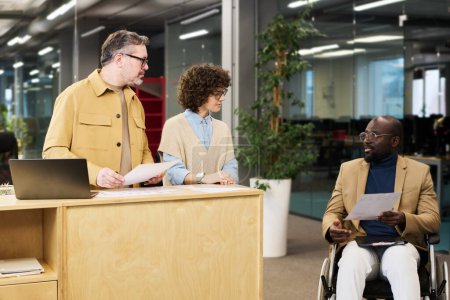 Téléchargez les photos : Des collègues confiants regardant l'analyste afro-américain handicapé assis en fauteuil roulant et consultant sur les données graphiques dans le document - en image libre de droit