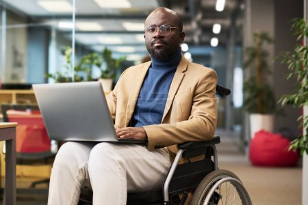 Jeune homme d'affaires afro-américain sérieux avec un ordinateur portable sur les genoux assis en fauteuil roulant dans un bureau ouvert et regardant la caméra