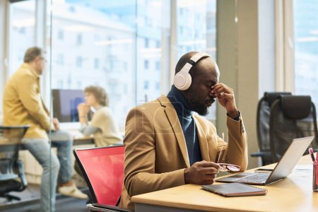 Téléchargez les photos : Jeune homme d'affaires fatigué dans des écouteurs assis sur le lieu de travail devant un ordinateur portable et ayant une courte pause contre ses collègues d'avoir parler - en image libre de droit