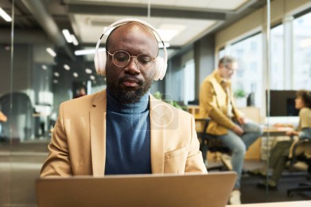 Téléchargez les photos : Jeune homme d'affaires afro-américain sérieux dans les écouteurs regarder en ligne vidéo sur les fluctuations financières à la bourse - en image libre de droit
