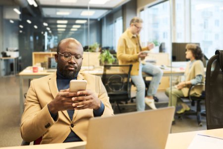 Photo for Serious African American manager using smartphone while sitting by workplace in front of laptop and texting against two colleagues - Royalty Free Image