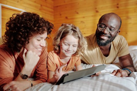 Téléchargez les photos : Heureux jeunes parents interculturels et leur mignon petit fils en utilisant une tablette tout en se reposant au lit le matin ensoleillé dans leur maison de campagne - en image libre de droit