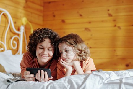Foto de Joven mujer sonriente con teléfono móvil y su hijo viendo películas o videos divertidos en línea mientras se relaja en la cama doble por la mañana - Imagen libre de derechos