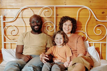 Foto de Alegre familia joven de hijo pequeño, su padre y su madre en ropa de casualrelax en la cama doble en casa y mirando a la cámara - Imagen libre de derechos