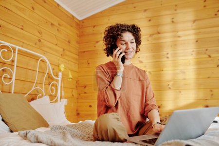 Foto de Joven mujer sonriente en pijama hablando en el teléfono móvil y la creación de redes delante de la computadora portátil mientras está sentado en la cama en casa - Imagen libre de derechos