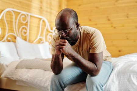 Téléchargez les photos : Jeune homme noir déprimé en tenue décontractée gardant les mains serrées par son visage tout en étant assis sur le lit le matin et en pensant à quelque chose - en image libre de droit