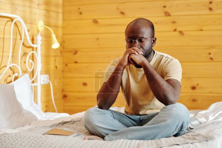 Téléchargez les photos : Jeune homme tendu en jeans et t-shirt gardant les mains serrées par son visage tout en étant assis sur le lit avec les jambes croisées et pensant - en image libre de droit