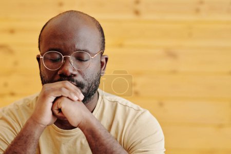 Téléchargez les photos : Jeune homme noir aux lunettes gardant les yeux fermés et les mains serrées par la bouche tout en se concentrant sur la résolution de problèmes graves - en image libre de droit