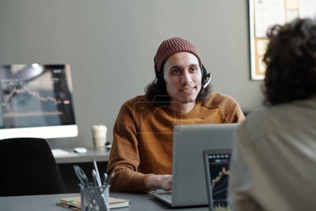 Foto de Joven hombre de negocios con auriculares mirando a una colega durante la comunicación en la oficina mientras ambos están sentados frente a las computadoras portátiles - Imagen libre de derechos