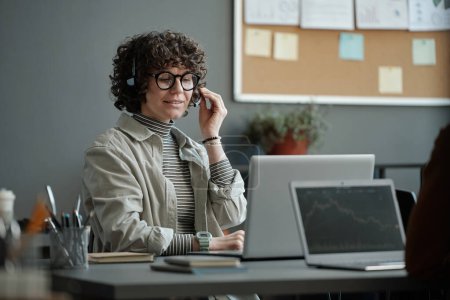 Foto de Joven operadora femenina confiada en auriculares sentada en el lugar de trabajo y hablando con el cliente en línea y ayudando a resolver problemas técnicos - Imagen libre de derechos