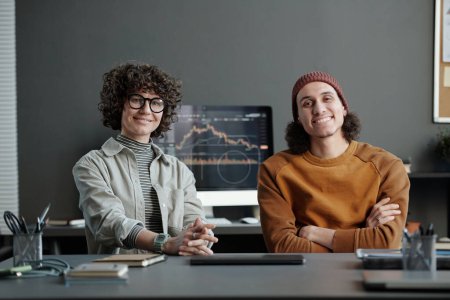 Téléchargez les photos : Deux jeunes responsables interculturels en tenue décontractée assis par lieu de travail au bureau et regardant caméra contre écran d'ordinateur avec graphique - en image libre de droit